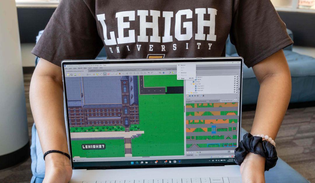 A student in a Lehigh University shirt holds a laptop showing a 2D game map editor.