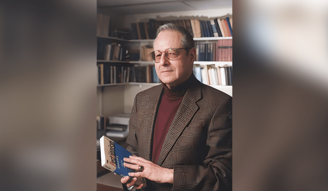 Photograph of an older man with glasses holding a book in front of bookshelves.