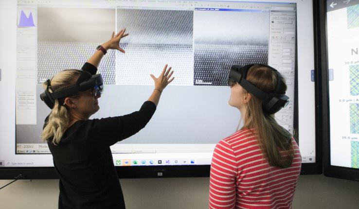 Two women wear VR headsets in front of a large touchscreen