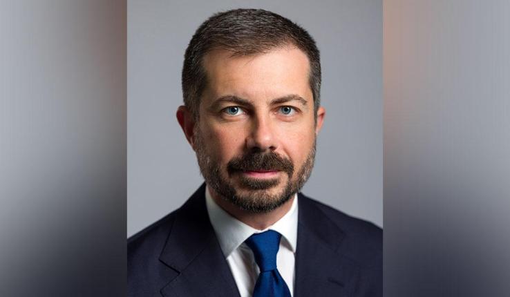 Professional portrait of Pete Buttigieg with a beard, wearing a blue tie and dark suit.