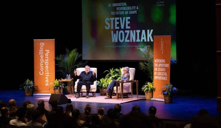 Steve Wozniak speaking on stage with audience, surrounded by plants and banners.