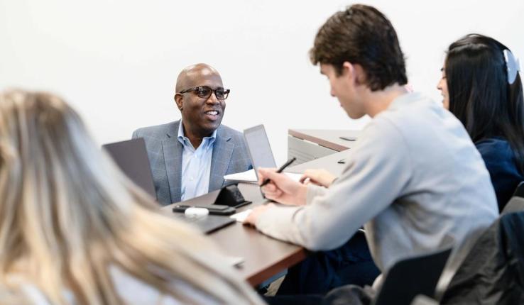 A professor sitting with students, all engaged in a discussion around a conference table.
