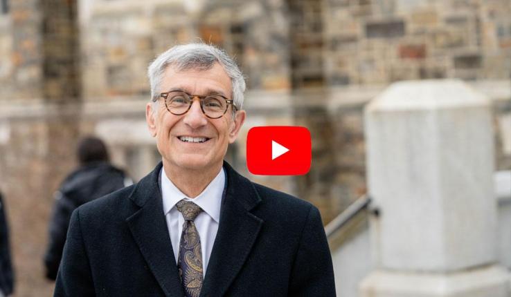 Smiling man in a black coat stands outside a stone building, with a YouTube logo in the corner.