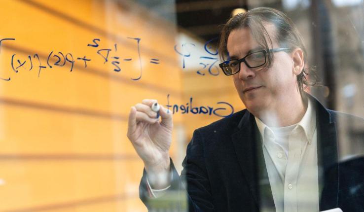 Man in glasses writing a complex math equation on a clear glass board.