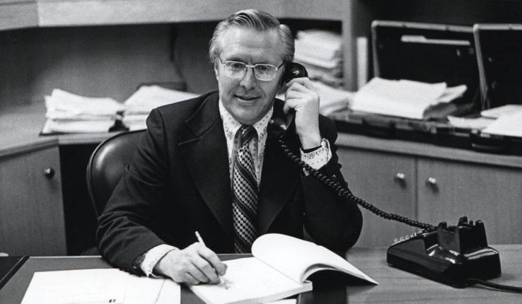 Black and white photograph: Man in suit and glasses talks on phone, writes at office desk.