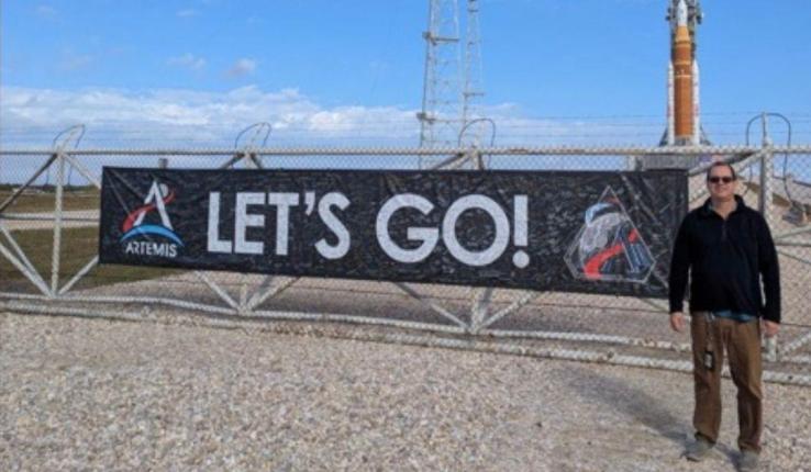 Photograph Mark Riddle by an Artemis "LET'S GO!" banner with a rocket in the background.