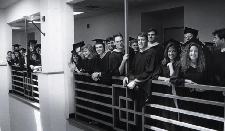 Black and white photograph of smiling graduates in caps and gowns along a balcony.