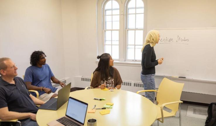 Four people collaborate in a bright classroom. One writes on a whiteboard, three watch from a table.