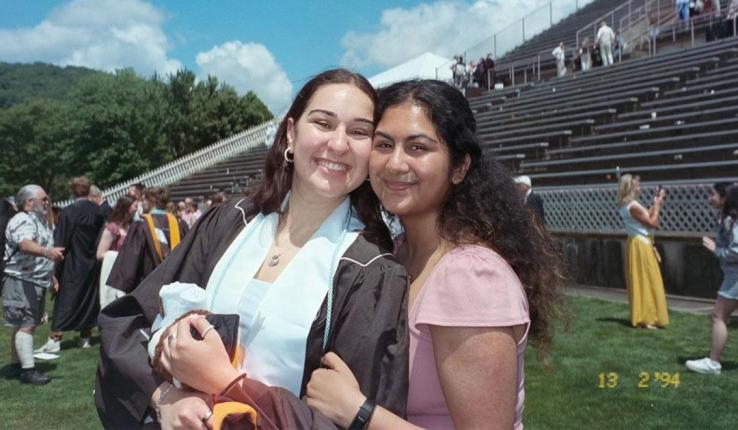 Photograph of two smiling women, one in a graduation gown, hugging at an outdoor ceremony.