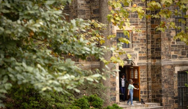 Photograph of people exiting a stone collegiate building framed by foliage.