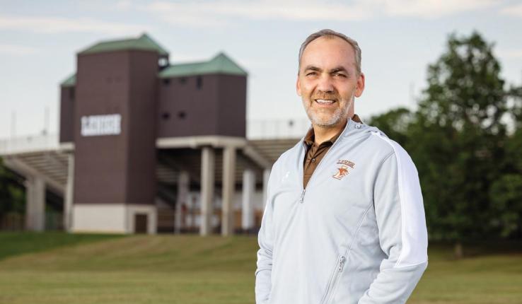 Smiling man in a grey athletic jacket with a sports stadium in the background.