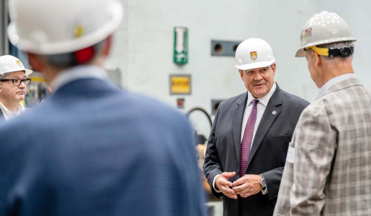 Photograph of a man in a suit and hard hat speaking to others in an industrial setting.