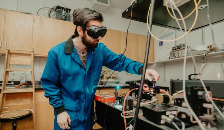 Photograph of a man in a blue lab coat and goggles working in a laboratory.